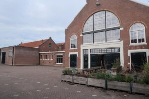 Vooraanzicht van het voormalige fabriekspand aan het Gedempt Hamerkanaal met grote ramen, zwarte deuren en een betegeld plein met plantenbakken.